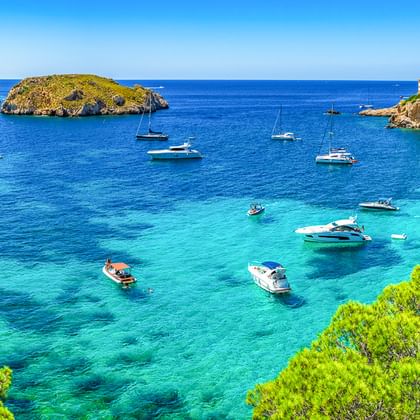 Panoramic view of Santa Ponça bay in Mallorca with turquoise water, boats anchored, rocky islands, and coastal buildings on cliffs framed by pine trees.