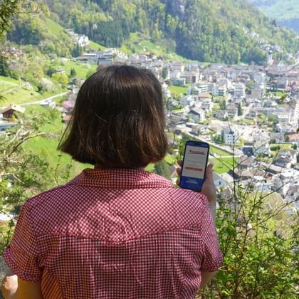 Frau im karierten Hemd hält Smartphone mit Eurotrek-App und blickt auf Alpendorf am See mit grünen Hügeln und Wäldern.