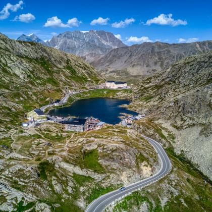 Luftaufnahme des Großen St. Bernhard Passes mit kurvenreicher Bergstraße, dunkelblauem Bergsee und Gebäuden im Tal mit schneebedeckten Gipfeln.