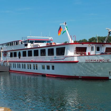 White river cruise ship MS Normandie with red trim anchored in calm blue water. German flag visible on stern, green trees in background.