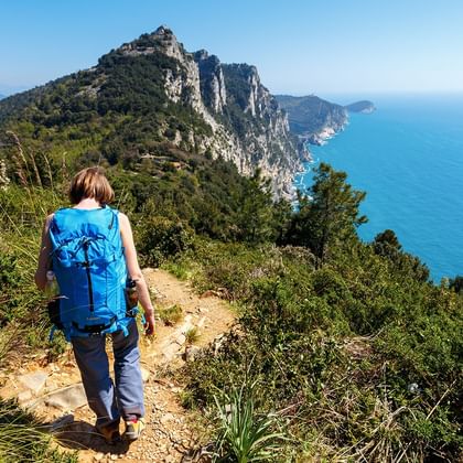 Wanderer mit blauem Rucksack auf Bergpfad in Cinque Terre mit Blick auf türkisfarbenes Mittelmeer und dramatische Felsenküste.