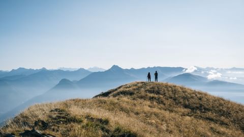 Zwei Wanderer auf einem grasbewachsenen Berggipfel im Tessin mit gestaffelten Bergketten und nebligen Tälern unter blauem Himmel.