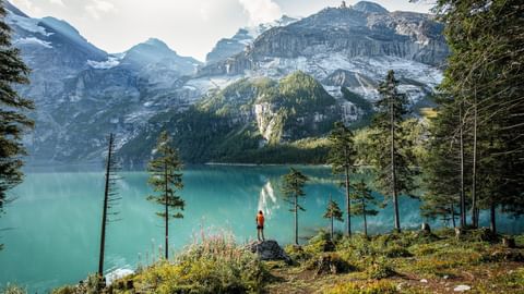 Wanderer in roter Jacke steht auf Felsvorsprung mit Blick auf den strahlend türkisen Oeschinensee, umgeben von schneebedeckten Alpengipfeln.