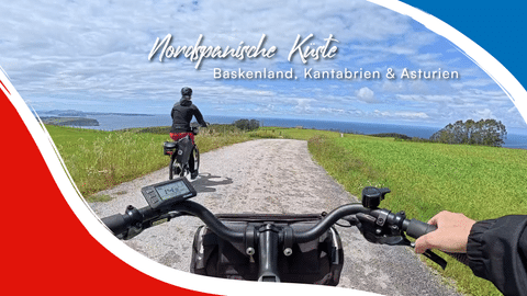 Blick vom Fahrradlenker auf Küstenweg in Nordspanien. Ein weiterer Radfahrer fährt voraus auf Schotterweg zwischen grünen Feldern und Meer.