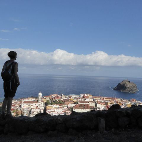Eine Wanderin blickt im Schatten hinunter auf die Stadt Garachico. Dahinter das Meer, in dem auf der rechten Seite ein grosser Fels aus dem Wasser ragt.