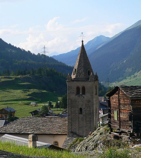 Rechts hat es ein kleines Holzhüttchen und gleich daneben der Glockenturm einer alten Kirche. Im Hintergrund ein Bergpanorama mit kleinen Wolken.