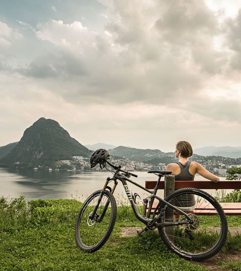 Une femme est assise sur un banc avec vue sur le Lago di Lugano et le San Salvatore.