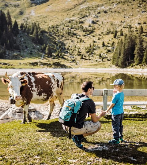 Ein Mann und sein Sohn stehen neben einer Kuh an einem See im Gantrisch Naturpark. 