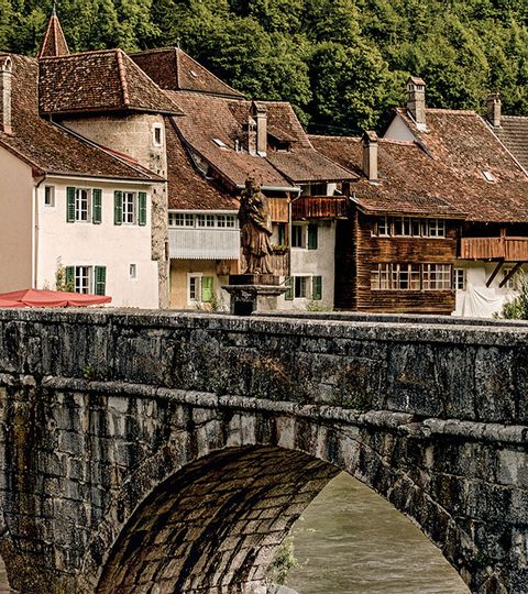 Ein Panoramabild das eine steinerene Bogenbrücke zu Saint Ursanne zeigt.