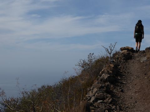 Eine Wanderin läuft einen schmalen Natusteinweg entlang. Links von ihr geht es bergabwärt und vor ihr Nebel und graue Wolken.