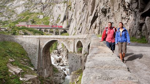 Zwei Wanderer wandern an einer Steinmauer entlang, im Hintergrund fährt ein Zug