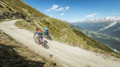 Zwei Mountainbiker auf einer Naturstrasse in den Bergen. Rechts eine Berglandschaft mit strahlend blauem Himmel.