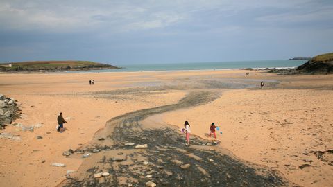 Die Harlyn-Bay. Ein riesen Sandstrand an dem Kinder spielen und verschiedene Menschen spazieren. Hinten links im Bild sieht ma noch einen kleinen Grashügel mit einem Häuschen und am Horizont ein bedekter grauer Himmel.