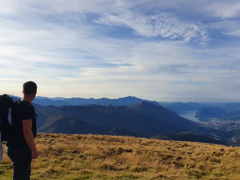 Wanderer schaut von einer Bergwiese rüber zum bewölkten, aber blauen Himmel über den Bergspitzen.