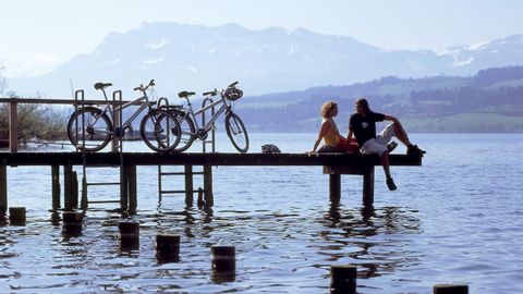 Two bikers take a break with their feet dangling. The bikes are leaning against the railing of the footbridge, with the mountains faintly visible in the background.