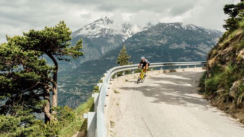 Cyclist coming out of a bend in a mountain landscape with snow-covered peaks.