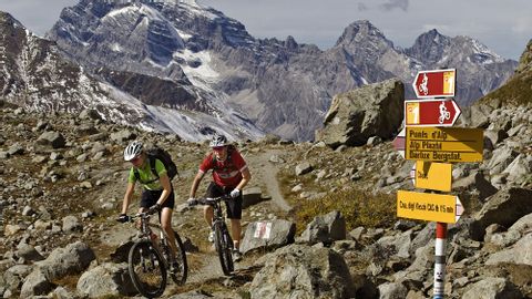Two mountain bikers in the Alps. A signpost shows them the route to Kesch in the Grisons mountains.