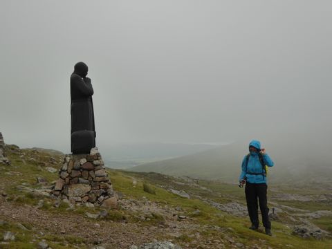 Eine Wanderin steht neben einer denkenden Statue unter grauem, nassem Wolkenhimmel.