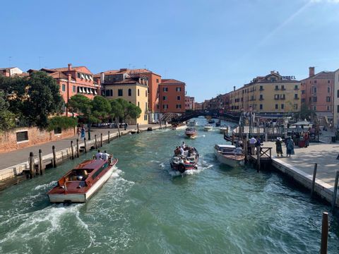 Man blickt auf einen Kanal in Venedig, auf dem viele Boote fahren. Farbige Häuser sind im Hintergrund zu sehen. 