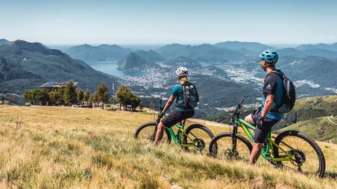 Un couple de VTTistes fait une pause dans le pâturage et regarde Lugano et les montagnes environnantes de loin.