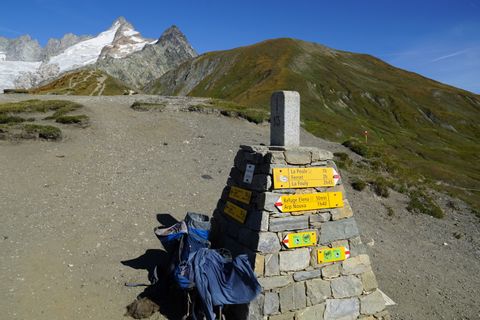 Vorne ein blauer Rucksack der an einem Grenzstein angelehnt ist, welcher auch Gleich als Wegweiser genutzt wird. Im Hintergrund die Spitze des mit Schnee bedeckten Mont-Blanc