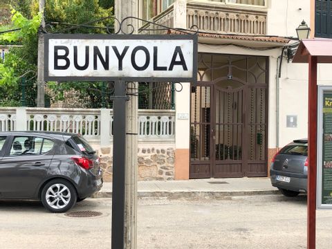 Ein weisses Schild auf der Srasse auf dem mit Grossbuchstaben in schwarz Bunyola steht. Hinter ihm ist ein Hauseingang der ein Tor aus Rostfarbenen Eisenstäben hat. auf der Strasse stehen 2 Autos von denen aber nur die Hecks zu sehen sind.