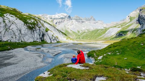 Mann und Kind sitzen auf einem Stein am Bach und blicken auf die Berglandschaft.