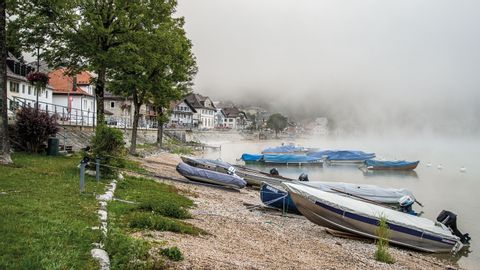 Le Pont im Nebel. Am steinigen Ufer des Sees warten einige kleine Motorboote auf ihre nächste fahrt.