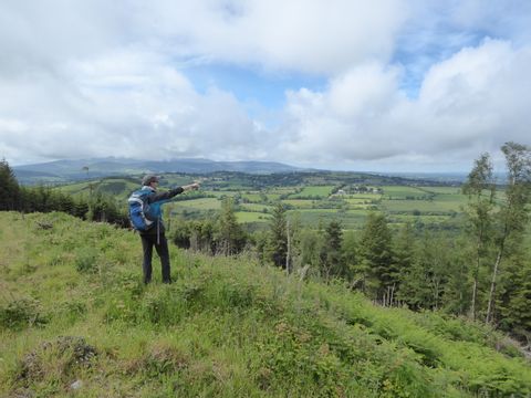 Auf dem grasigen Hügel steht ein Wanderer, der in die weite, grüne Landschaft zeigt, die tiefer unten ist. Der Himmel ist hellblau aber mit vielen Cumulus Wolken bedeckt.