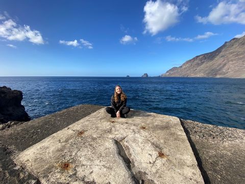 Riana sitzt auf einem Stein und in ihrem Hintergrund liegt die Küste von El Hierro. 