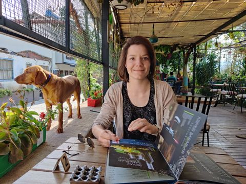 Eine Frau sitzt am Tisch in einem Restaurant, links von ihr ein Hund. Im Hintergrund sind Pflanzen zu sehen. 