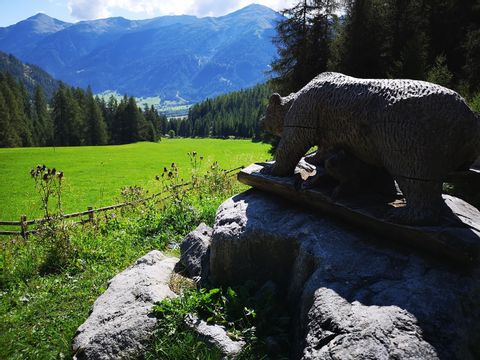 Bärenstatue auf einem Grossen Stein welcher vor einer Idylischen Berg und Wiesenlandschaftsteht. 