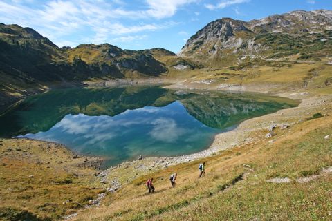 Trois randonneurs au lac de Formarine sur le Lechweg. Un lac de montagne bleu avec un ciel bleu et un paysage de collines.
