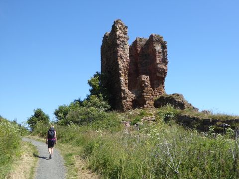 Wanderin spaziert auf einem schmalen Weg zwischen Wiesen. Im Hintergrund die Mauern einer Ruine.
