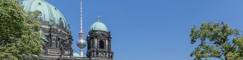 Auf einem Panoramabild sind rechts ein paar Baumäste mit sattgrünen Blätter. Rechts ist der Berliner Funkturm zwischen einer grossen und einer kleinen Kirchenkuppel unter strahlend blauem Sommerhimmel zu sehen.