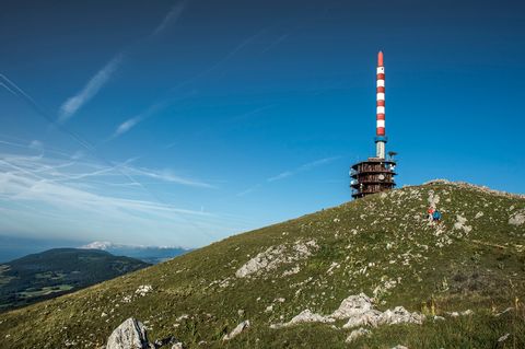 Auf einer Bergspitze steht ein Rot-Weiss gestreifter Funkmast vor einem strahlend blauen Himmel. Ganz klein versteckt sieht man 2 Wandere die den Berg hinunter laufen.