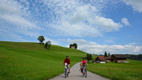 A couple is cycling on a road between pastures. On the right in the background is a farm with an attached barn.