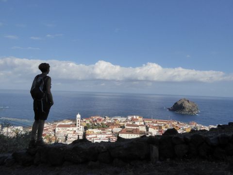 Eine Wanderin blickt im Schatten hinunter auf die Stadt Garachico. Dahinter das Meer, in dem auf der rechten Seite ein grosser Fels aus dem Wasser ragt.