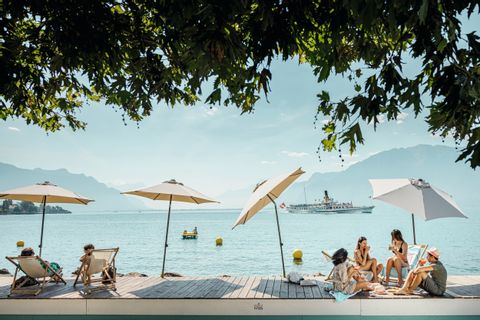 Ein paar Sonnenschirme auf einem Steg, im Hintergrund ein Schiff auf dem Genfersee. 