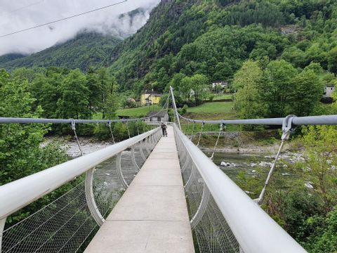 Man sieht wie eine Velofahrerin die Hängebrücke bei Maggia überquert. Im Hintergrund ist in Mitten von Bäumen ein kleines Dorf erkennbar. 