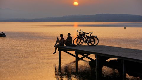 Two bikers sit on the jetty with their bikes, watching a beautiful sunset.