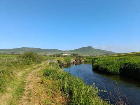 Path beside a river in Dingle, Ireland, in bright sunshine. Green hills in the background. 