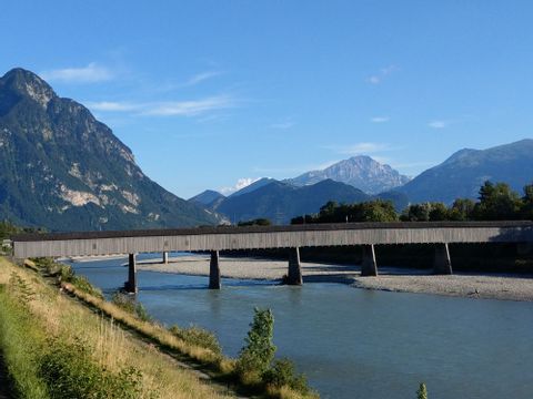 Eine Holzbrücke führt über einen Fluss mit Bergen im Hintergrund. 