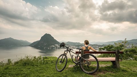 A woman sits on a bench with a view of Lago di Lugano and San Salvatore.