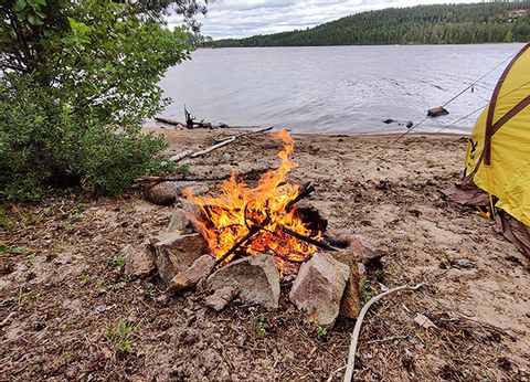 Feuer vor einem See mit Zelt im vordergrund.