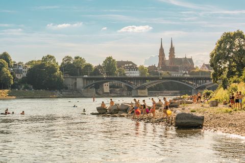 Menschen gehen über eine Sandbank in den Rhein schwimmen. Im Hintergrund das Basler Münster. 