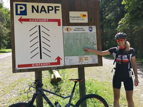 Eine Velofahrerin zeigt auf die grosse Holztafel. Zu sehen ist die Luzerner vWanderwegkarte und die Aufteilung des Parkplatzes Napf.