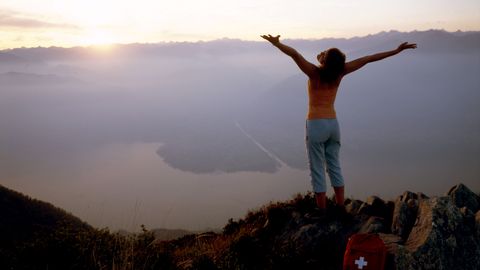 Frau mit ausgebreiteten Armen auf einem Berggipfel, die den Sonnenaufgang hinter dem See begrüsst