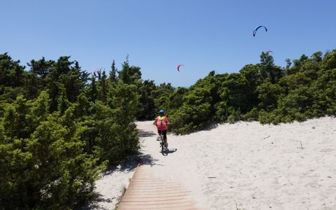 Radfahrerin fährt auf einem Weg am Sandstrand mit Gleitschirmfliegern im Hintergrund