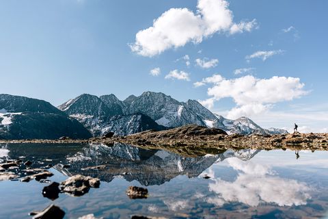 A mountain backdrop is reflected in a mountain lake. A hiker in the background. 